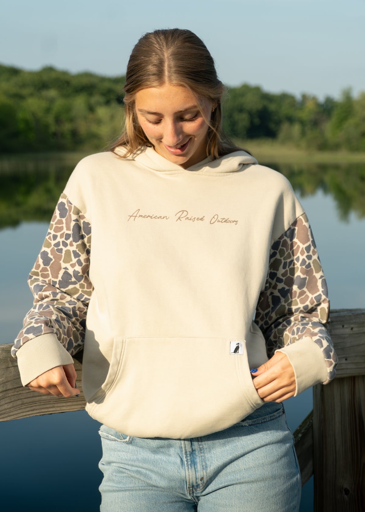 women wearing oversized tan hoodie with camo sleeves by a lake